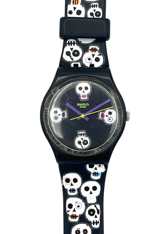 A Swatch wristwatch with a black case and a black strap decorated with a repeating pattern of white skulls with colorful eyes and teeth, set against a plain white background.