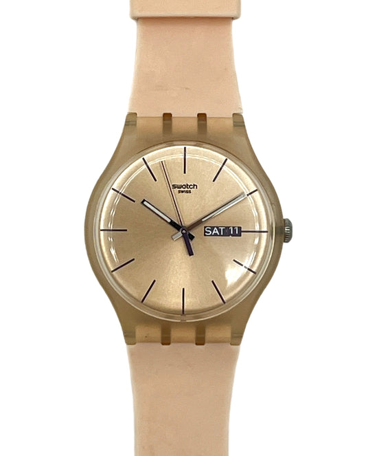 A Swatch wristwatch with a light beige silicone strap and a matching beige dial, housed in a bronze-toned case. The watch features black hands, simple hour markers, and a day/date window displaying 'SAT 11'.