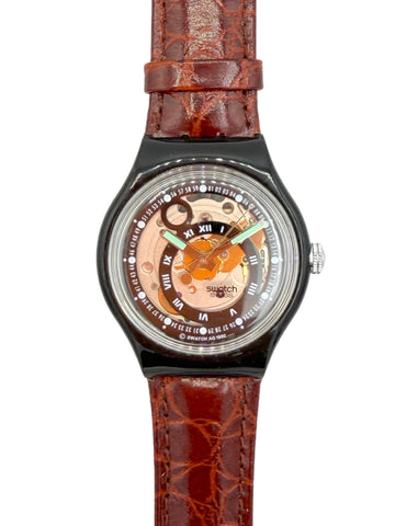 A wristwatch with a brown textured leather strap, a black bezel, and a transparent dial showing a gold-toned inner mechanism and Roman numeral hour markers.