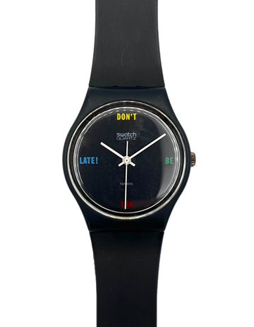 A Swatch wristwatch with a black silicone strap and a black round dial. The dial features white hands, a yellow 'DON'T' at the 12 o'clock position, and blue and green text at the 9 and 3 o'clock positions, with a small red 'LATE!' at the 6 o'clock position.